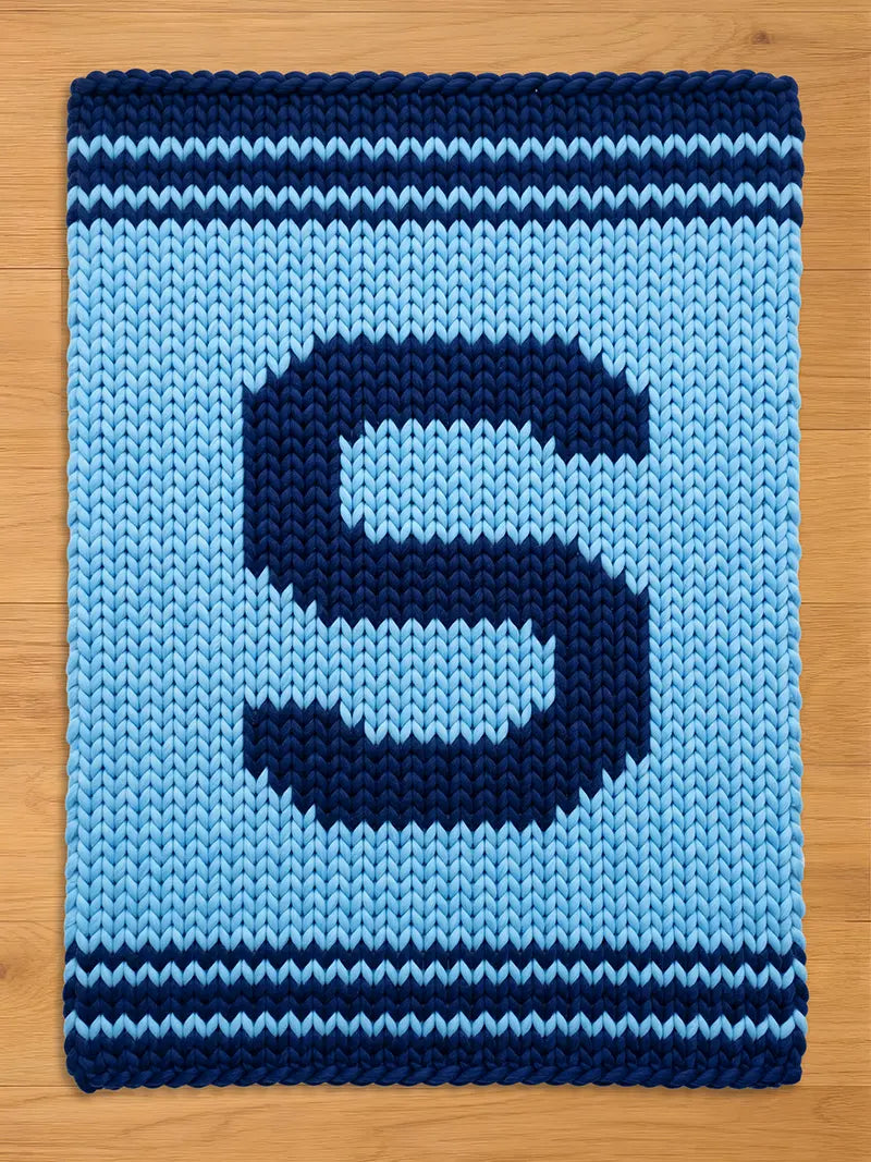 A knitted rectangular chunky blanket in shades of blue. The blanket features a large, dark blue letter "S" in the center, surrounded by a lighter blue background. Dark blue stripes border the top and bottom edges. 