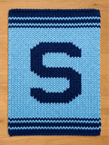 A knitted rectangular chunky blanket in shades of blue. The blanket features a large, dark blue letter "S" in the center, surrounded by a lighter blue background. Dark blue stripes border the top and bottom edges. 