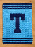 A knitted blanket in shades of blue, featuring a large, dark blue letter "T" in the center. The blanket has a textured appearance with horizontal stripes at the top and bottom. The background is a light blue color.