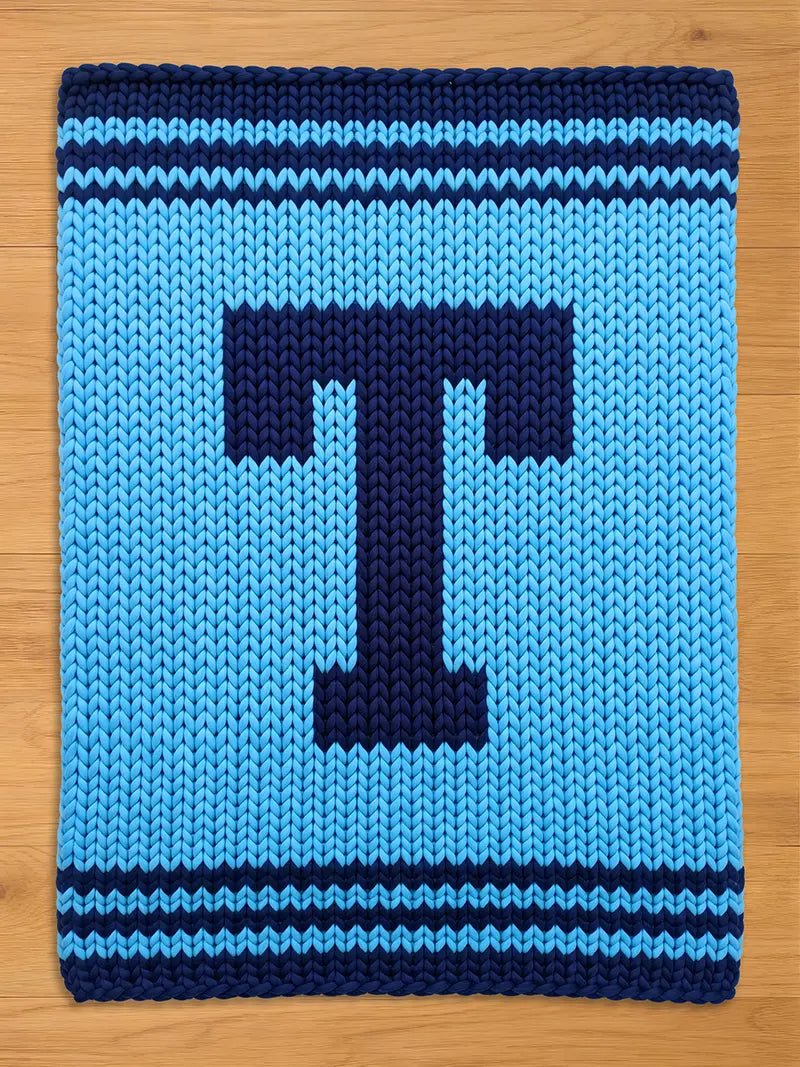 A knitted blanket in shades of blue, featuring a large, dark blue letter "T" in the center. The blanket has a textured appearance with horizontal stripes at the top and bottom. The background is a light blue color.