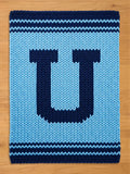A knitted blanket in shades of blue, featuring a large letter "U" in navy blue against a lighter blue background. The blanket has navy blue stripes at the top and bottom, creating a simple, yet bold design. The texture is chunky and cozy.