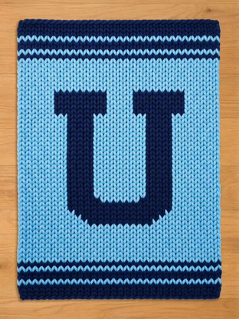 A knitted blanket in shades of blue, featuring a large letter "U" in navy blue against a lighter blue background. The blanket has navy blue stripes at the top and bottom, creating a simple, yet bold design. The texture is chunky and cozy.