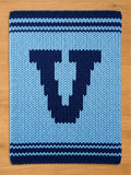 A knitted blanket in shades of blue, featuring a large, dark blue letter "U" in the center. The blanket has a light blue background with dark blue stripes at the top and bottom, and is made of thick yarn.