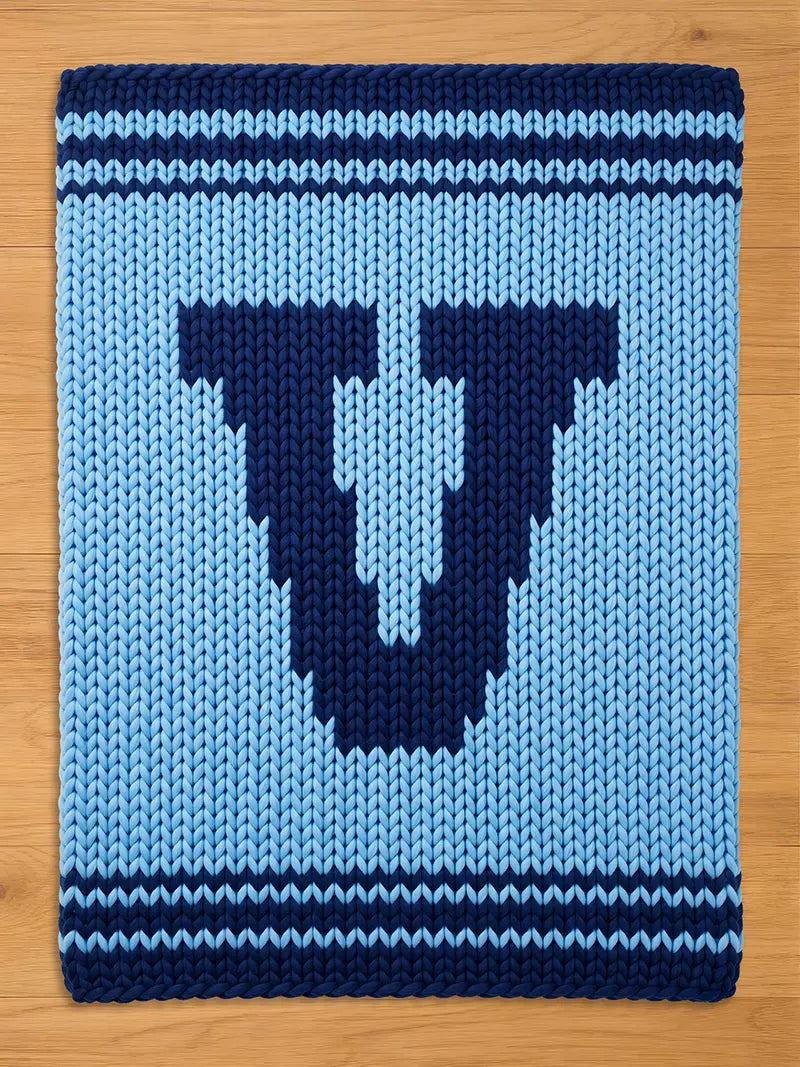 A knitted blanket in shades of blue, featuring a large, dark blue letter "U" in the center. The blanket has a light blue background with dark blue stripes at the top and bottom, and is made of thick yarn.