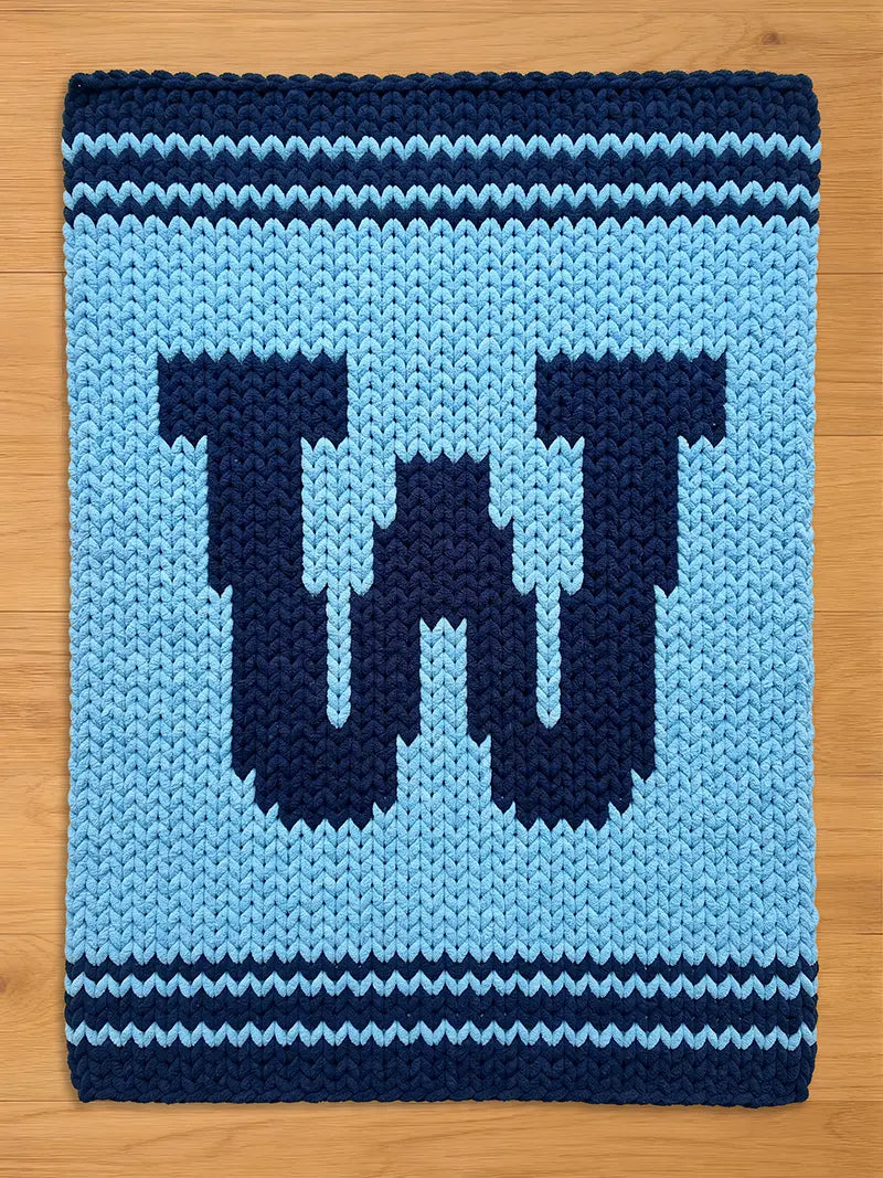 A knitted, light blue blanket with a dark blue letter "W" in the center. The blanket has dark blue stripes at the top and bottom, and is made of a soft, chunky yarn. The blanket is rectangular.