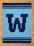 A knitted, light blue blanket with a dark blue letter "W" in the center. The blanket has dark blue stripes at the top and bottom, and is made of a soft, chunky yarn. The blanket is rectangular.