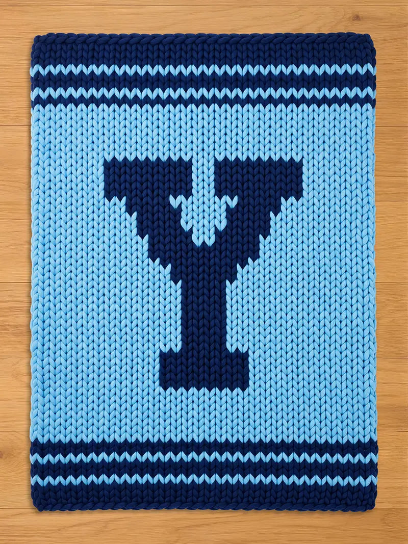 A knitted blanket in light blue with a dark blue letter "Y" in the center. The blanket has dark blue stripes at the top and bottom. The chunky knit design gives it a cozy, handmade feel.