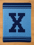 A knitted blanket in shades of blue, featuring a large, dark blue letter "X" in the center. The blanket has a light blue background and dark blue borders with horizontal stripes. The item is made of thick yarn.