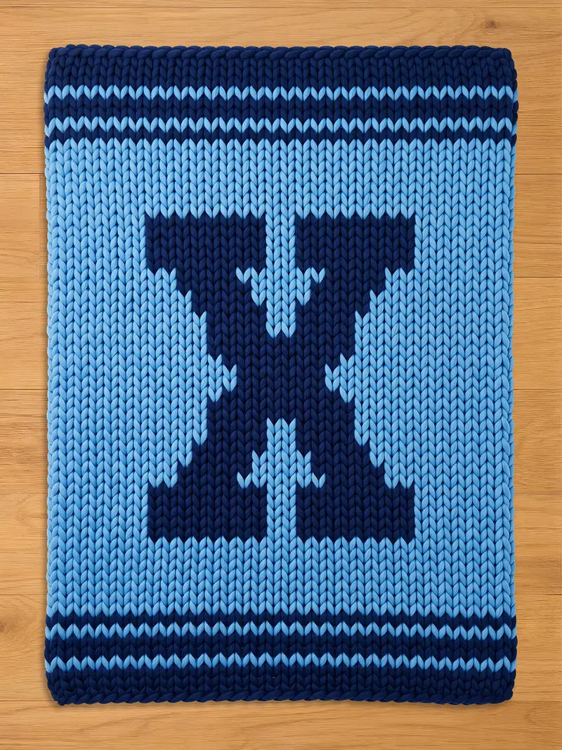 A knitted blanket in shades of blue, featuring a large, dark blue letter "X" in the center. The blanket has a light blue background and dark blue borders with horizontal stripes. The item is made of thick yarn.