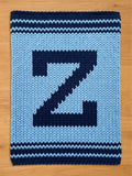 A knitted blanket featuring a large, dark blue letter "Z" against a light blue background. The blanket has dark blue stripes at the top and bottom edges. The chunky knit design gives it a cozy, handmade appearance