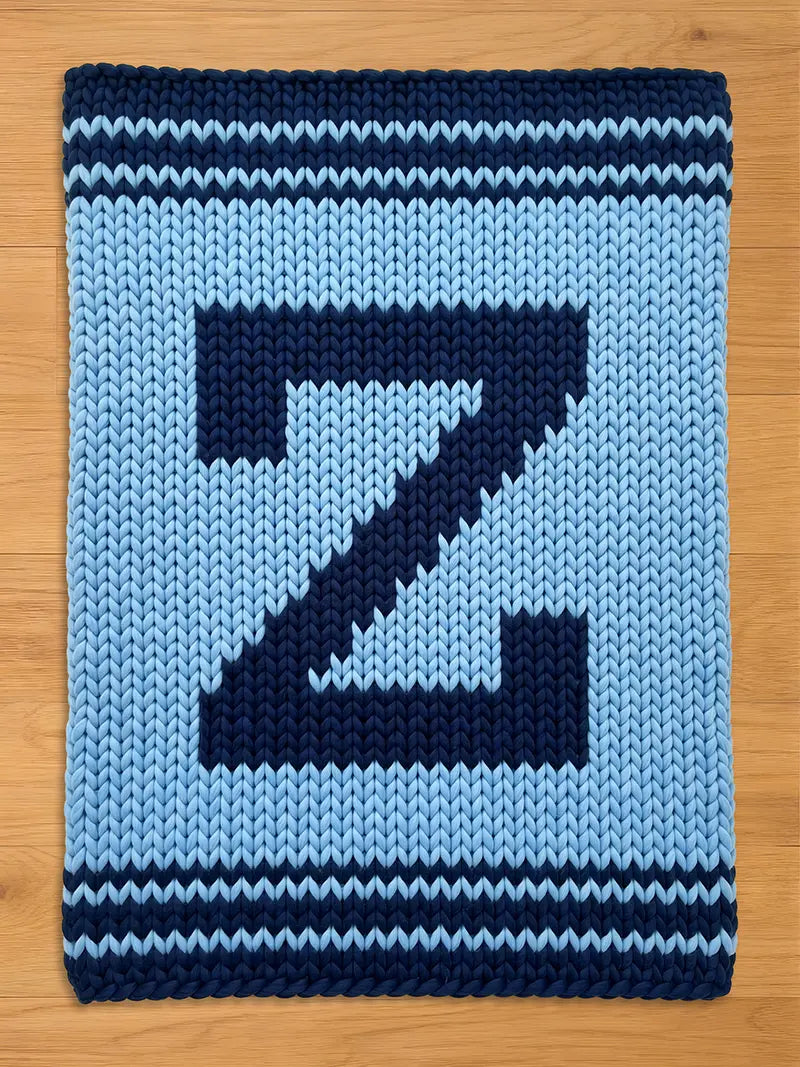 A knitted blanket featuring a large, dark blue letter "Z" against a light blue background. The blanket has dark blue stripes at the top and bottom edges. The chunky knit design gives it a cozy, handmade appearance