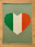 A knitted blanket featuring a heart-shaped design in the colors of the Irish flag: green, white, and orange. The blanket has a light green background and a textured appearance, suitable for home decor.