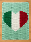 A knitted rectangular chunky blanket with a heart-shaped design in the colors of the Italian flag: green, white, and red. The background is a light green color. The blanket is made of thick yarn.
