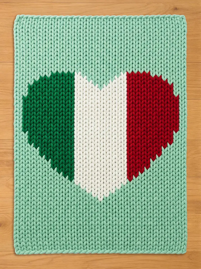 A knitted rectangular chunky blanket with a heart-shaped design in the colors of the Italian flag: green, white, and red. The background is a light green color. The blanket is made of thick yarn.