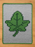 A rectangular, knitted chunky blanket featuring a large green leaf design on a gray background. The leaf is rendered in shades of green, with a darker green outline. The blanket has a green border.