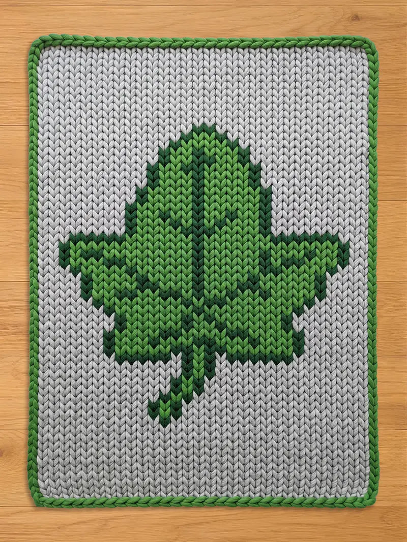A rectangular, knitted chunky blanket featuring a large green leaf design on a gray background. The leaf is rendered in shades of green, with a darker green outline. The blanket has a green border.