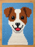 A knitted chunky blanket featuring a smiling dog face. The dog has brown ears, black eyes, and a white and brown face. The background is light blue, and the dog's mouth is pink. The square is made of yarn.