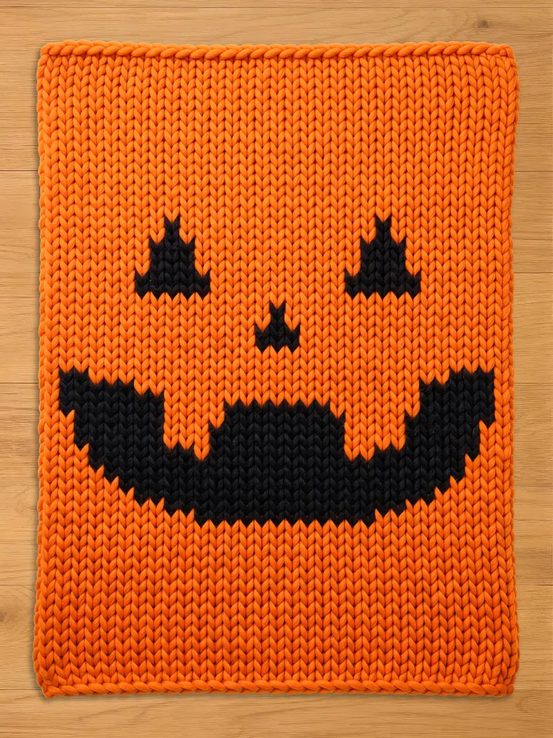A rectangular chunky knit blanket in bright orange featuring a black jack-o'-lantern face pattern. The design includes two triangular eyes, a small triangular nose, and a wide, jagged, toothy grin centered on the orange yarn.