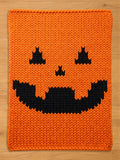 A rectangular chunky knit blanket in bright orange featuring a black jack-o'-lantern face pattern. The design includes two triangular eyes, a small triangular nose, and a wide, jagged, toothy grin centered on the orange yarn.
