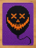 A rectangular knitted blanket featuring a purple background and a large black balloon shape in the center. The balloon has an orange stitched design of two X-shaped eyes and a wide, jagged, toothy grin, with a black string hanging down.