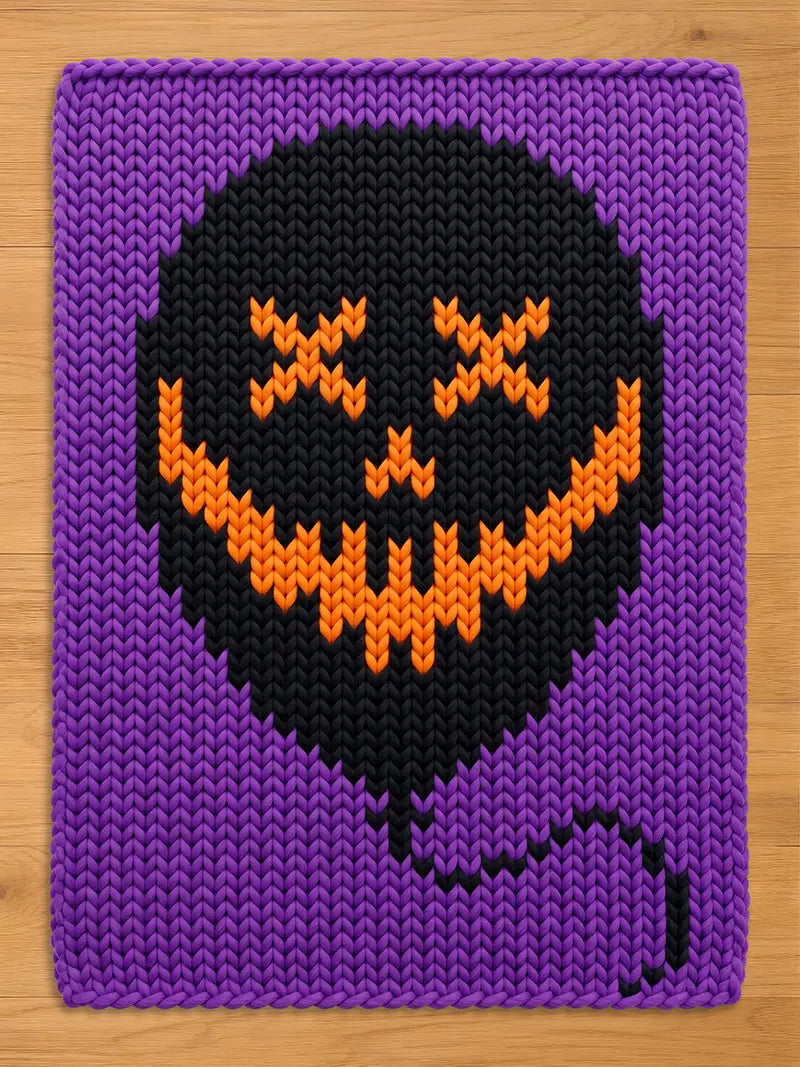 A rectangular knitted blanket featuring a purple background and a large black balloon shape in the center. The balloon has an orange stitched design of two X-shaped eyes and a wide, jagged, toothy grin, with a black string hanging down.