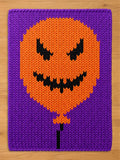 A rectangular knitted chunky blanket featuring a purple background and a central orange balloon shape. The balloon has a black knitted jack-o'-lantern face with jagged eyes and a wide, toothy grin, with a black string extending downward.