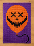 A rectangular knitted blanket featuring a large orange balloon with a black jack-o'-lantern face, including X-shaped eyes and a jagged mouth, set against a solid purple background with a black string extending from the bottom.