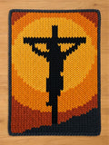 Chunky knit blanket featuring a black silhouette of Jesus on the cross against a warm yellow, orange, and red sunset background, finished with a dark grey knitted border