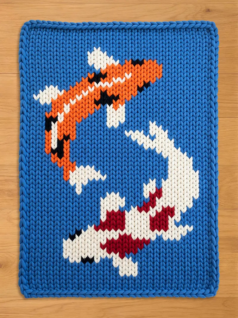 A knitted blue blanket featuring two koi fish in orange, white, black, and red. The fish are swimming in opposite directions against a blue background. The blanket has a thick, textured appearance.
