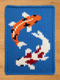 A knitted blue blanket featuring two koi fish in orange, white, black, and red. The fish are swimming in opposite directions against a blue background. The blanket has a thick, textured appearance.