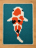 A knitted chunky blanket featuring a koi fish design. The fish is rendered in orange, white, and black against a teal background. The rug has a cream-colored border and is placed on a wooden surface.