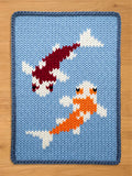 A knitted chunky blanket featuring two koi fish swimming on a light blue background. One fish is red and white, the other is orange and white. The border is a darker blue.
