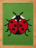 A rectangular knitted blanket featuring a large ladybug design centered on a bright green background. The ladybug has a vibrant red body with black spots, a black head with white accents, and black legs extending to the sides.