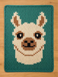 A chunky knit blanket in teal featuring a large, centered design of a cute llama face in cream, tan, and black yarn