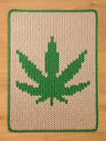A rectangular, knitted chunky blanket featuring a large green cannabis leaf design on a cream-colored background. The leaf is centrally placed, with a green border outlining the rug's edges. The blanket is made of thick yarn.