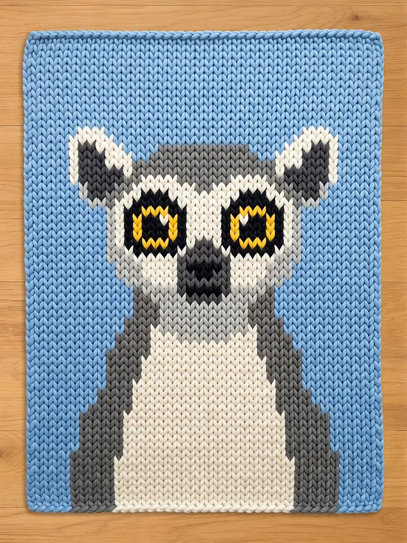 A knitted chunky blanket featuring a lemur design. The lemur has large yellow eyes, a black nose, and a white and gray body. The background is a light blue color. The knitted texture is visible throughout the design.