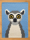 A knitted chunky blanket featuring a lemur design. The lemur has large yellow eyes, a black nose, and a white and gray body. The background is a light blue color. The knitted texture is visible throughout the design.
