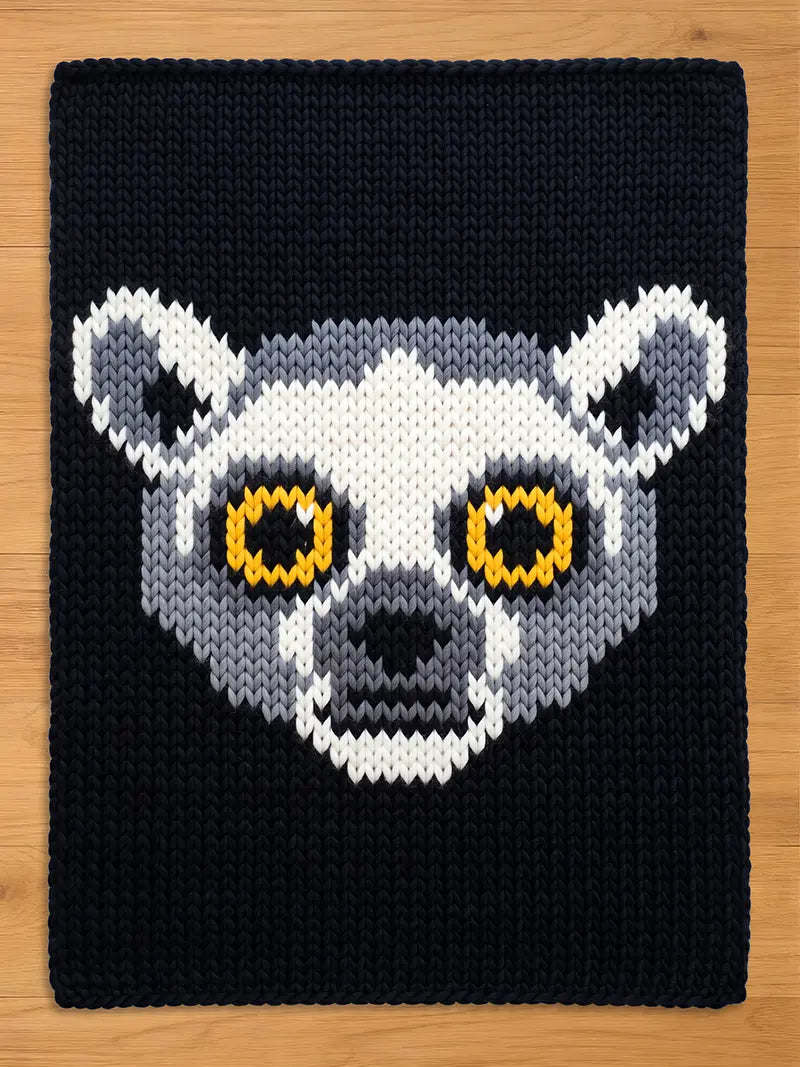 A knitted chunky blanket featuring a lemur face design. The lemur's face is rendered in white, gray, and black, with yellow eyes. The background is a solid navy blue.