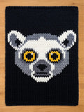 A knitted chunky blanket featuring a lemur face design. The lemur's face is rendered in white, gray, and black, with yellow eyes. The background is a solid navy blue.