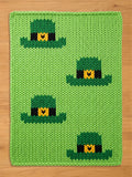 A green knitted rectangular chunky blanket featuring four green leprechaun hats with black bands and gold buckles. The background is a solid light green color. The blanket is made of a thick, textured yarn.