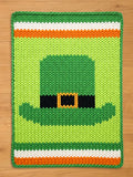 Chunky knit blanket featuring a central green leprechaun hat with a black band and orange buckle, set against a light green background with green, white, and orange stripes at the top and bottom