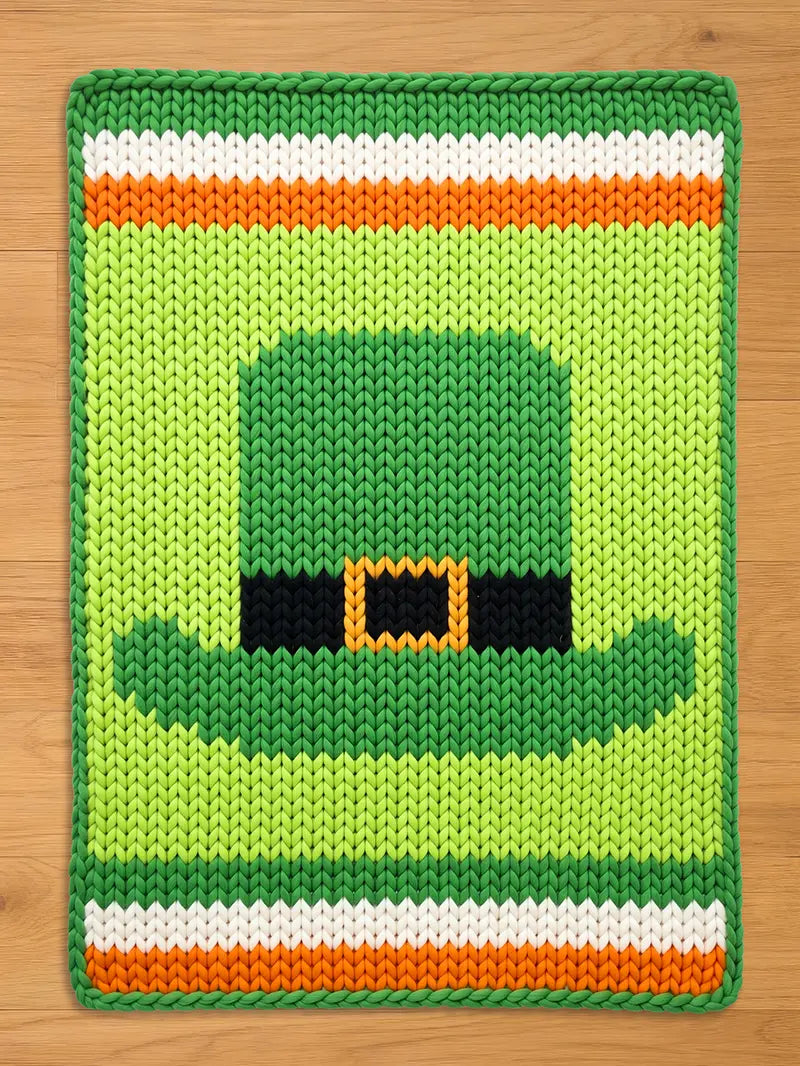 Chunky knit blanket featuring a central green leprechaun hat with a black band and orange buckle, set against a light green background with green, white, and orange stripes at the top and bottom