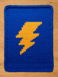 Knitted blanket in a vibrant blue with a large yellow lightning bolt design. The blanket is made of thick yarn, creating a textured appearance. The bold colors and graphic design make it a striking home decor item.