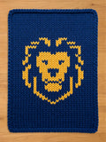 A dark blue chunky knit blanket laid flat on a light wooden floor, featuring a large, stylized golden-yellow lion head pattern knitted into the center