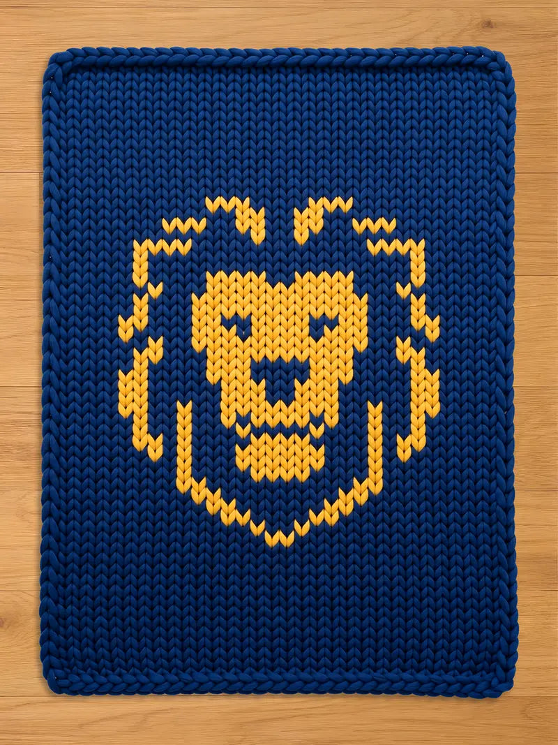A dark blue chunky knit blanket laid flat on a light wooden floor, featuring a large, stylized golden-yellow lion head pattern knitted into the center
