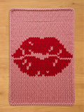 A knitted pink chunky blanket with a large red lip design. The lips have white accents. The blanket is rectangular with a knitted border and is placed on a wooden surface.