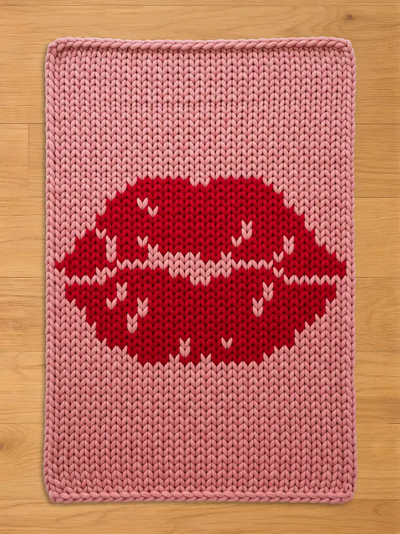A knitted pink chunky blanket with a large red lip design. The lips have white accents. The blanket is rectangular with a knitted border and is placed on a wooden surface.