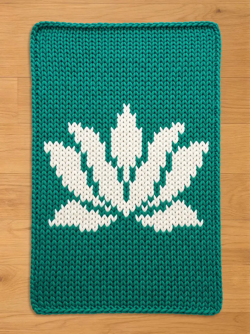 A rectangular, knitted teal-colored chunky blanket with a white lotus flower design. The lotus flower is centered on the mat and is made of white yarn. The blanket is made of thick yarn and has a textured appearance.
