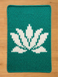 A rectangular, knitted teal-colored chunky blanket with a white lotus flower design. The lotus flower is centered on the mat and is made of white yarn. The blanket is made of thick yarn and has a textured appearance.