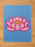 Rectangular chunky knit blanket with a light blue background, featuring a large, central lotus flower design in light pink with dark pink outlines and a yellow core, laid flat on a wooden floor.